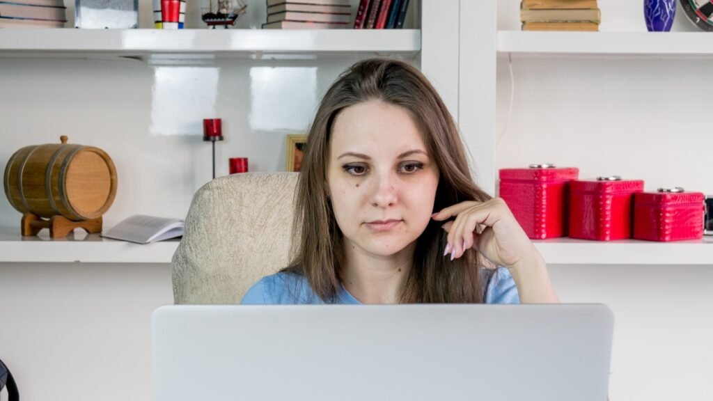 A woman sitting at a laptop in a home office, resting her chin on her hand with an unfocused, distracted expression.