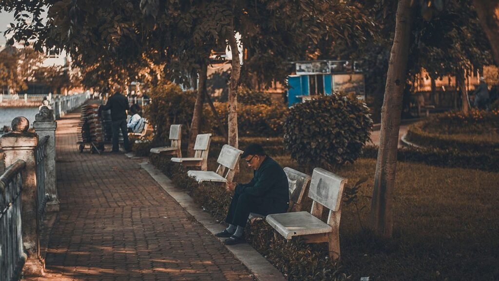 An older man sitting alone on a park bench at golden hour, with other people further along the tree-lined path behind him.