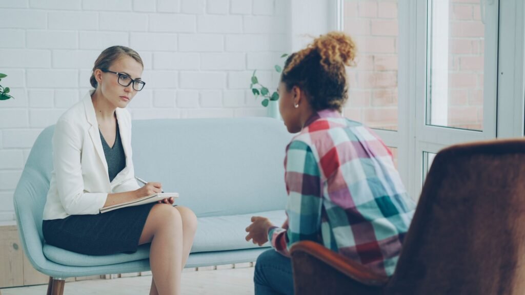 A therapist with a blank notepad listening attentively to a client during a counseling session in a bright, plant-filled room