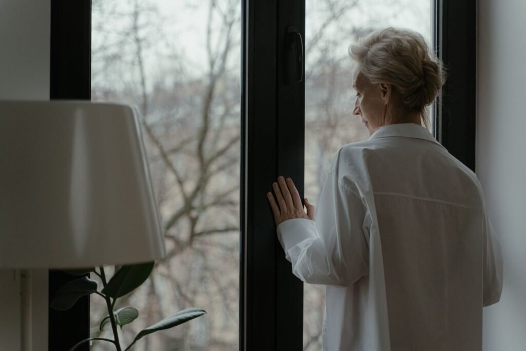 An older woman in a white shirt stands at a window with her hand resting on the frame, looking out at bare winter trees.