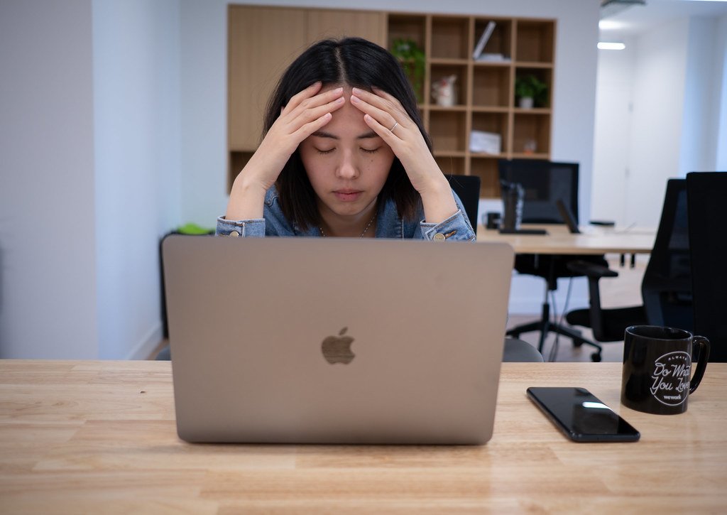 A woman sits at a desk with her laptop, eyes closed and head in her hands, showing visible exhaustion and worry.