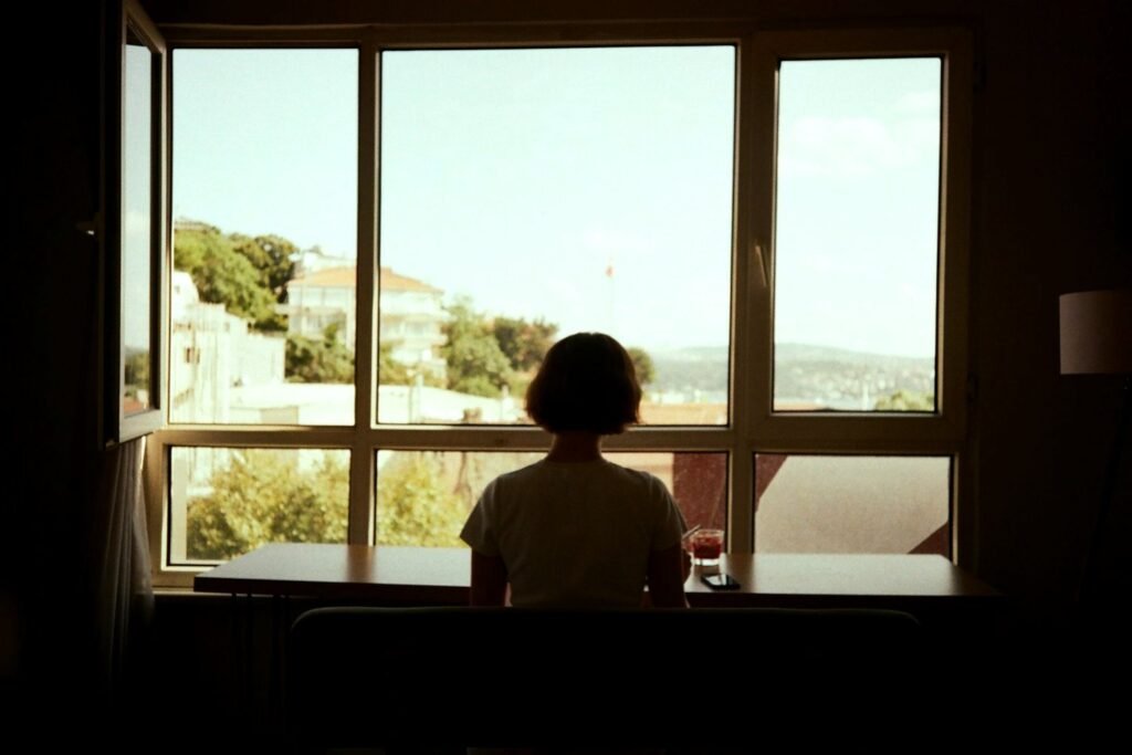 A person sitting alone at a table by a large open window, seen from behind, looking out at daylight over rooftops and trees.