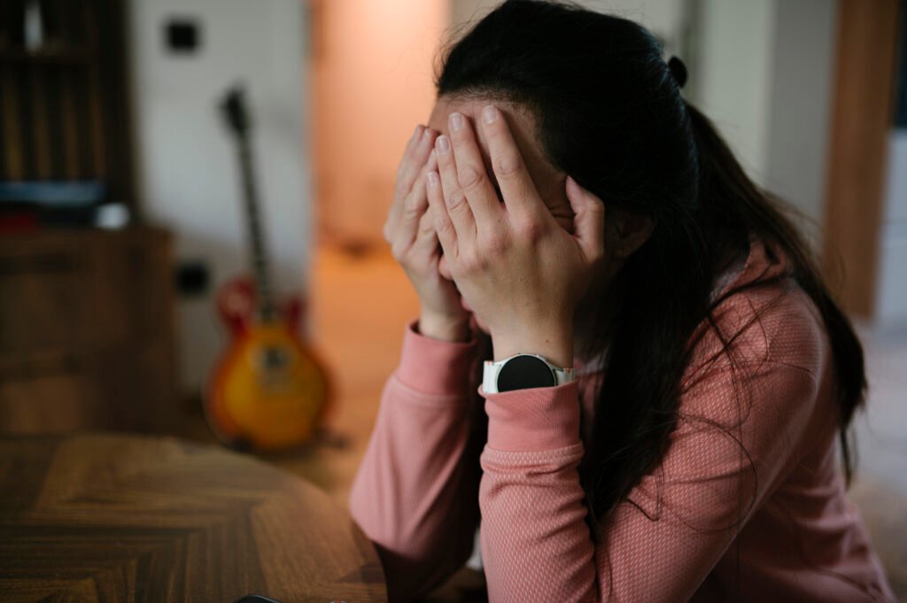 A woman sits at a table with her face in her hands, taking a moment to calm her breathing during a stressful moment at home.