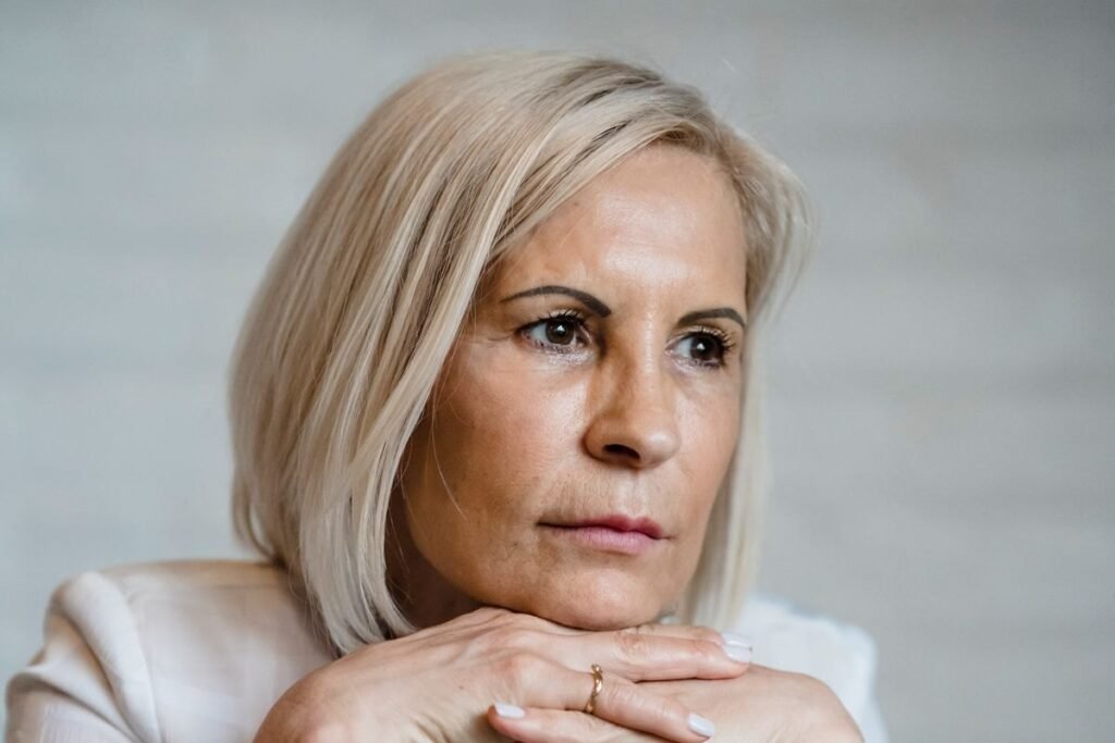 Thoughtful woman in her late forties resting her chin on her hands, looking away from the camera in a quiet, brightly lit room