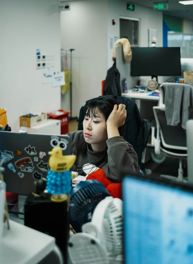 A woman sits at her office desk in the evening, resting her head tiredly on her hand while looking at her laptop screen.
