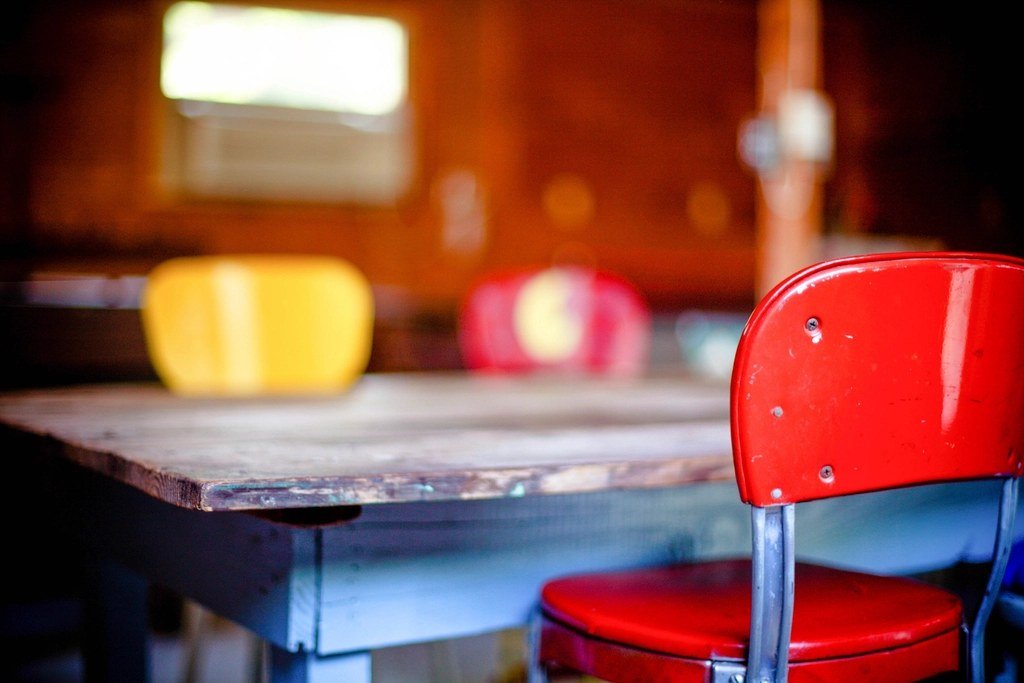 An empty red chair pulled up to a worn wooden desk in a quiet, warmly lit classroom, with other empty seats blurred in the background.