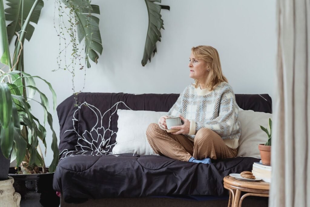 A woman in her fifties sits cross-legged on a sofa in a plant-filled living room, holding a mug and gazing thoughtfully out of frame.