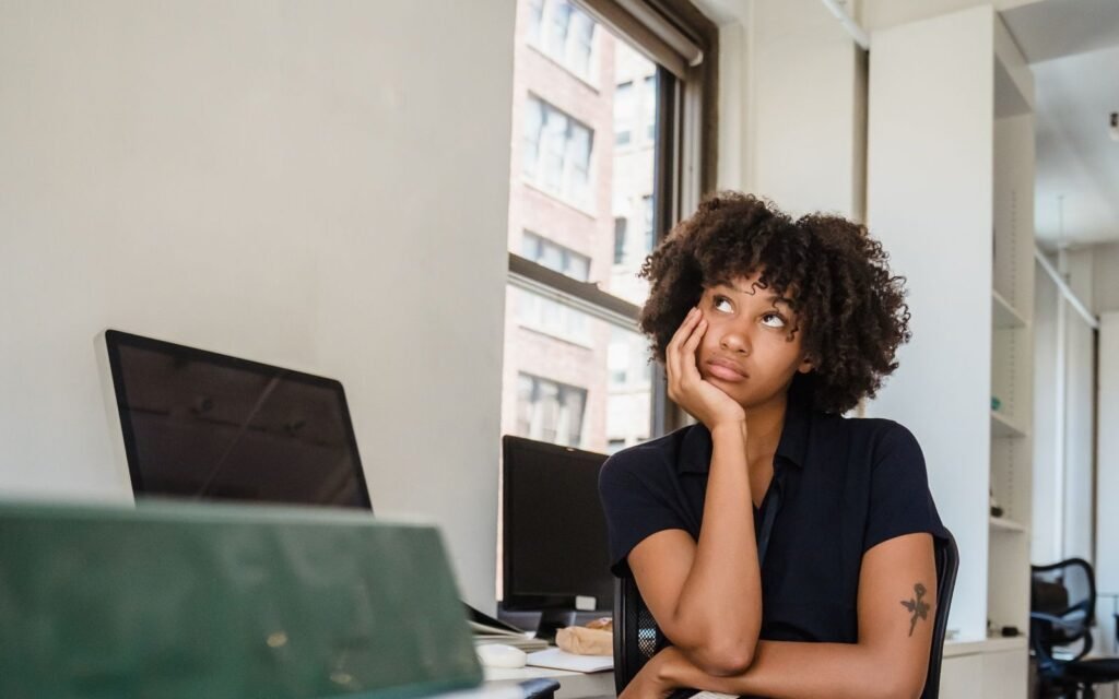 A young woman with curly hair sits at an office desk beside a window, chin resting on her hand, gazing upward with a distant expression while monitors and binders sit in front of her.