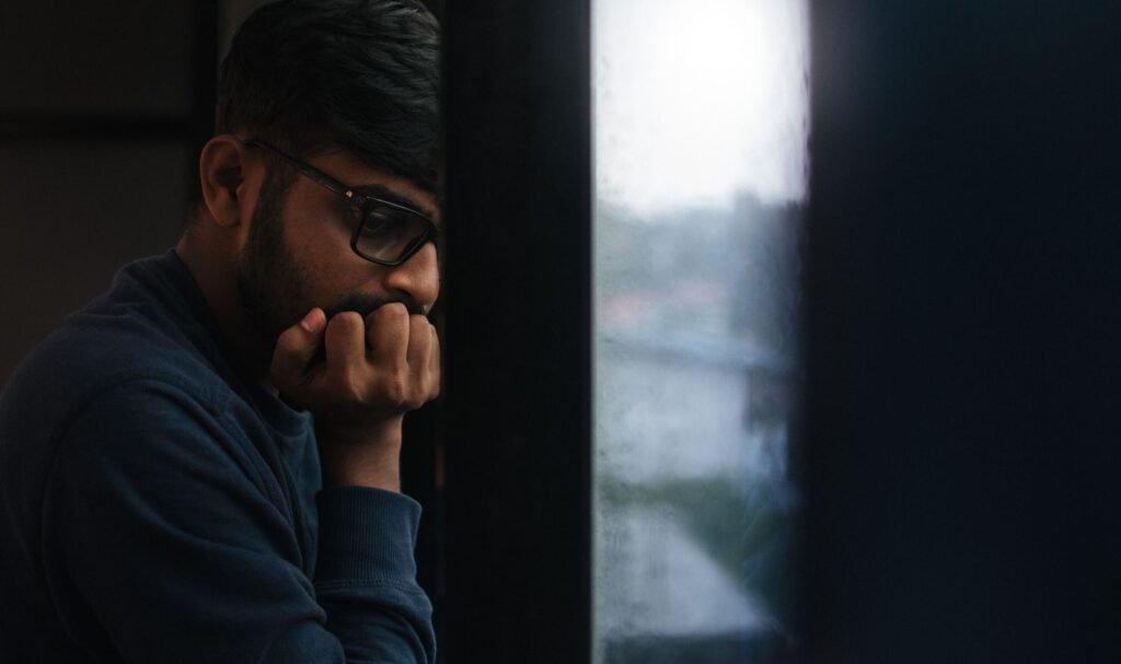 A man in glasses rests his chin on his hand, gazing out a rain-streaked window, lost in thought.